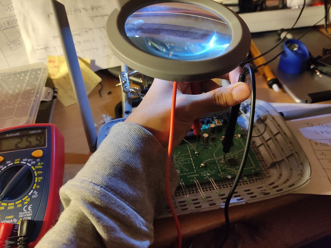 Probing the router board with an AstroAI multimeter under a magnifier lamp, the handwritten pinout notebook visible at right